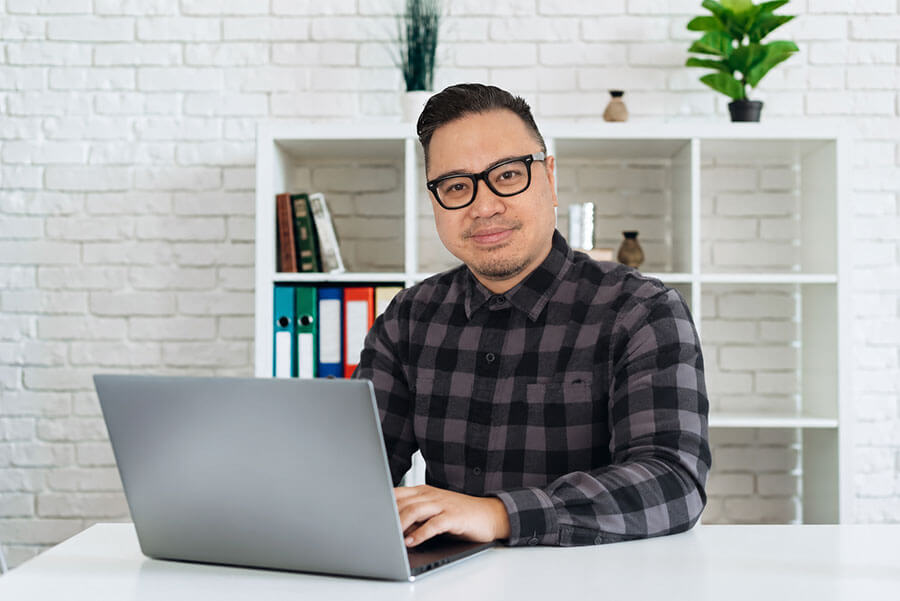 Nick Tolentino, web designer and creative lead for Piranha Studios sitting at a desk on a laptop. The desk is white and the background is a white brick background with a half empty bookshelf with a couple of green plants on top.